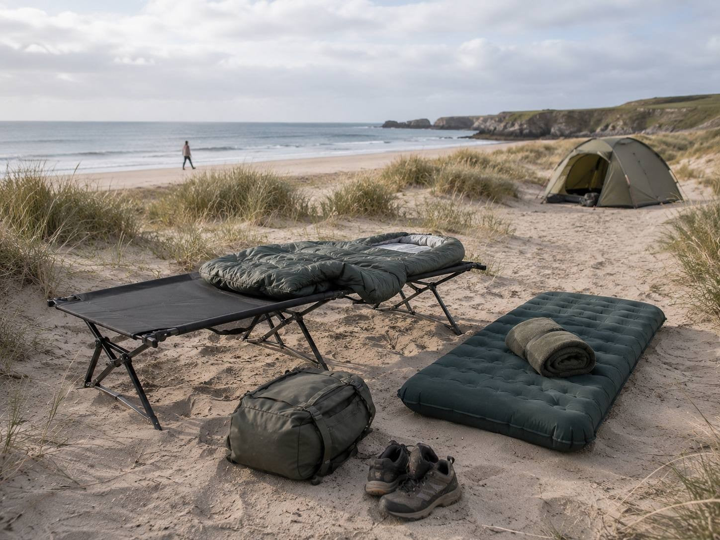 A camping setup on sand dunes overlooking the ocean, featuring a black folding camp cot with a green sleeping bag, a sleeping pad, a duffel bag, and hiking boots, with a tent in the background.