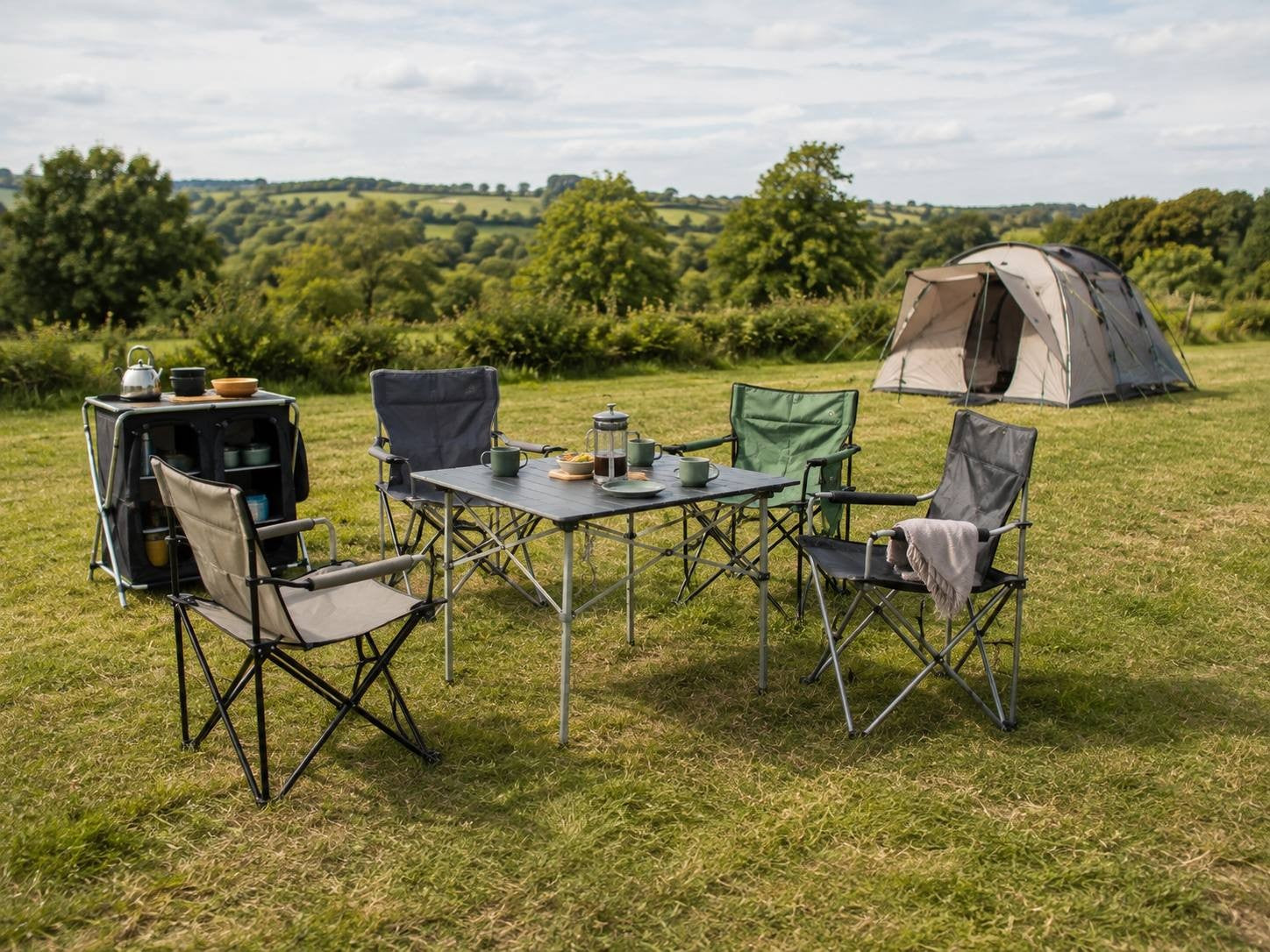 An outdoor campsite setup on a green meadow featuring a folding camping table surrounded by various folding chairs, a portable kitchen storage unit with a kettle, and a family tent in the background under a cloudy sky.