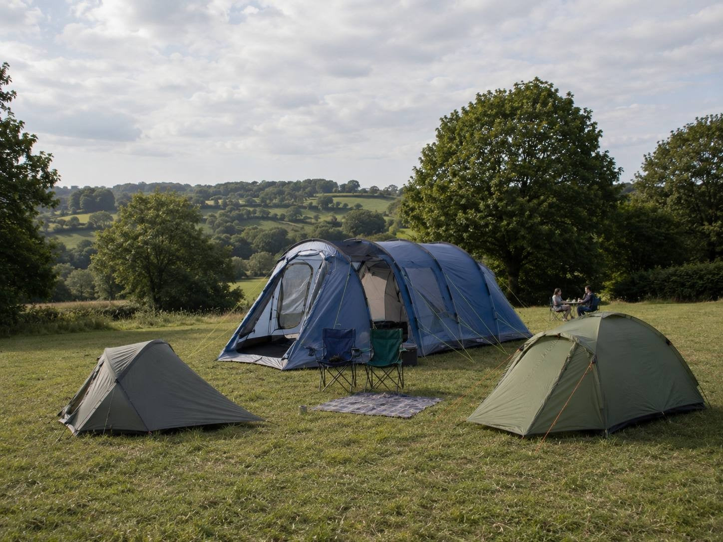 A scenic campsite on a grassy field featuring a large blue multi-room family tent in the center, flanked by two smaller green dome tents and folding chairs under a cloudy sky.