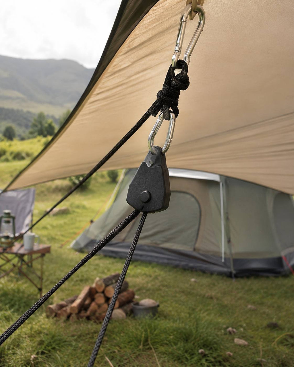 A close-up shot of a black rope ratchet attached to a beige camping tarp and tied to a secure anchor point near a tent in a grassy field.