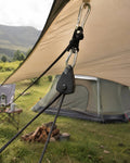 A close-up shot of a black rope ratchet attached to a beige camping tarp and tied to a secure anchor point near a tent in a grassy field.