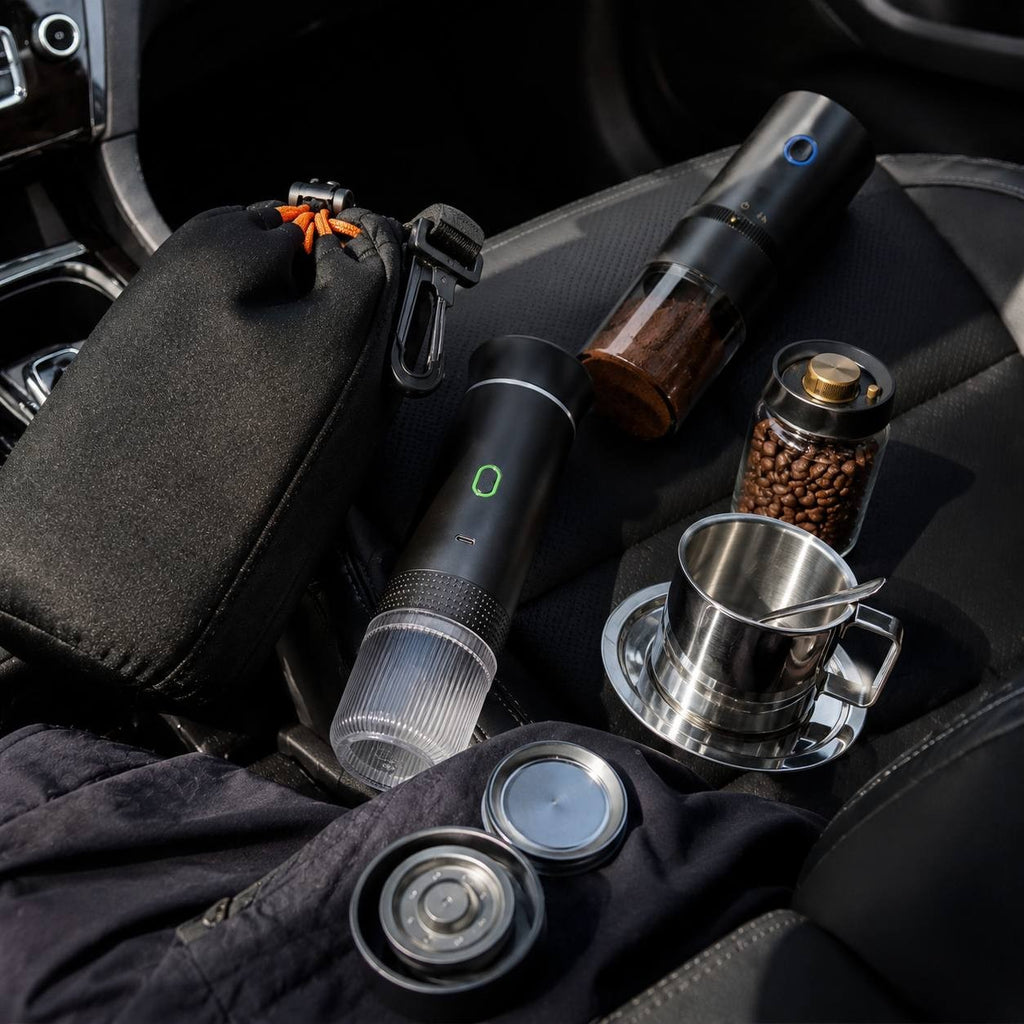 Disassembled travel coffee set on a dark leather car seat including a black carry pouch bag, espresso maker, automatic grinder, and stainless steel cup.