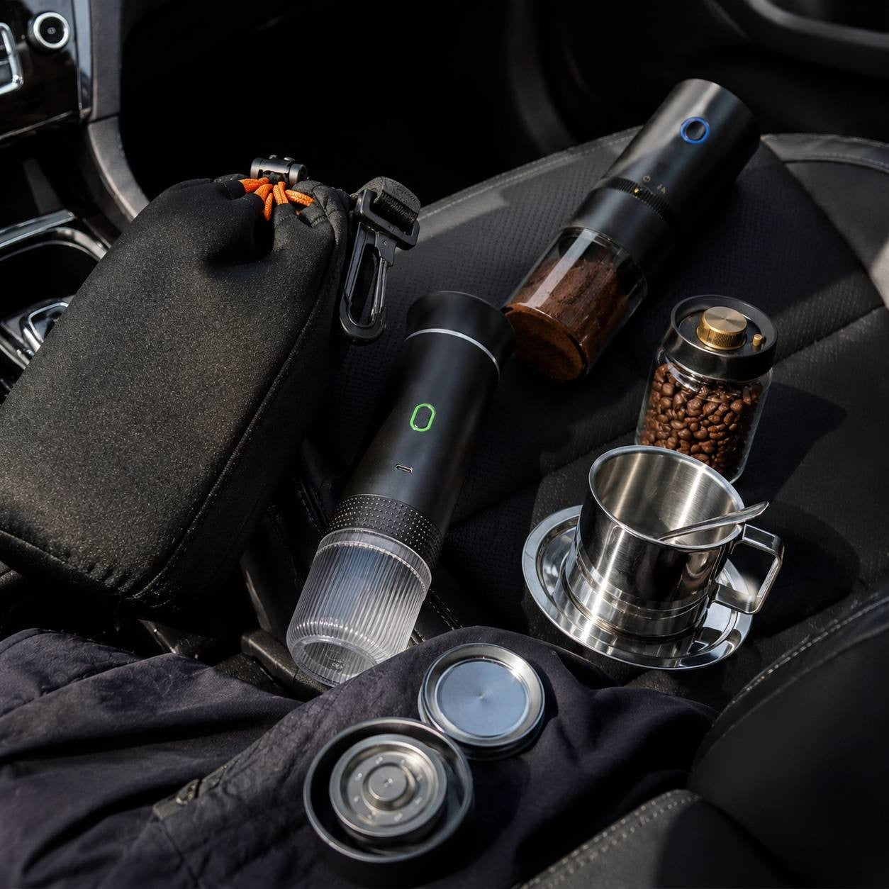 Disassembled travel coffee set on a dark leather car seat including a black carry pouch bag, espresso maker, automatic grinder, and stainless steel cup.