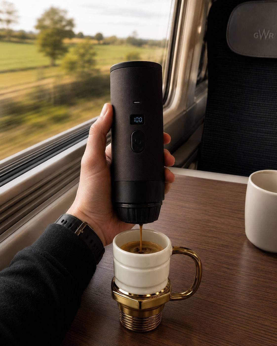 Close-up of a hand inside a train carriage (GWR) holding a portable coffee maker, which is pouring coffee into a white ceramic mug with a distinct gold-colored spark plug base and handle, placed on a wooden table.