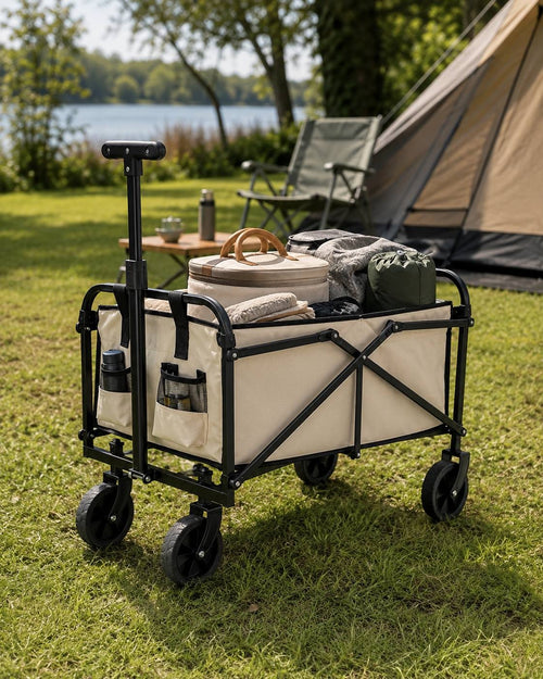 Beige collapsible utility wagon loaded with bags and camping gear, set up on grass at a campsite with a lake and forest in the background.