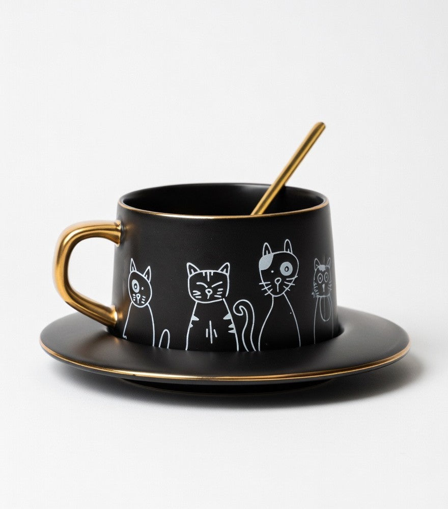 Black mug with gold handle and saucer featuring white cat illustrations on a white background