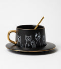 Black mug with gold handle and saucer featuring white cat illustrations on a white background