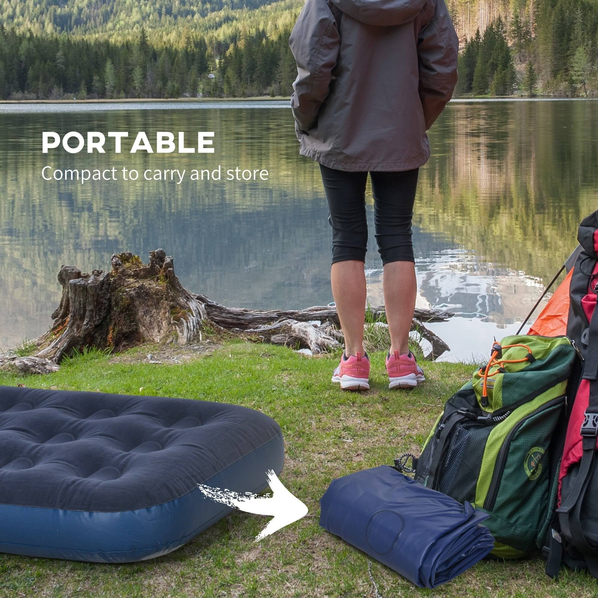 Rolled-up blue air mattress placed on the grass next to backpacks near a lake with a person standing in the background.