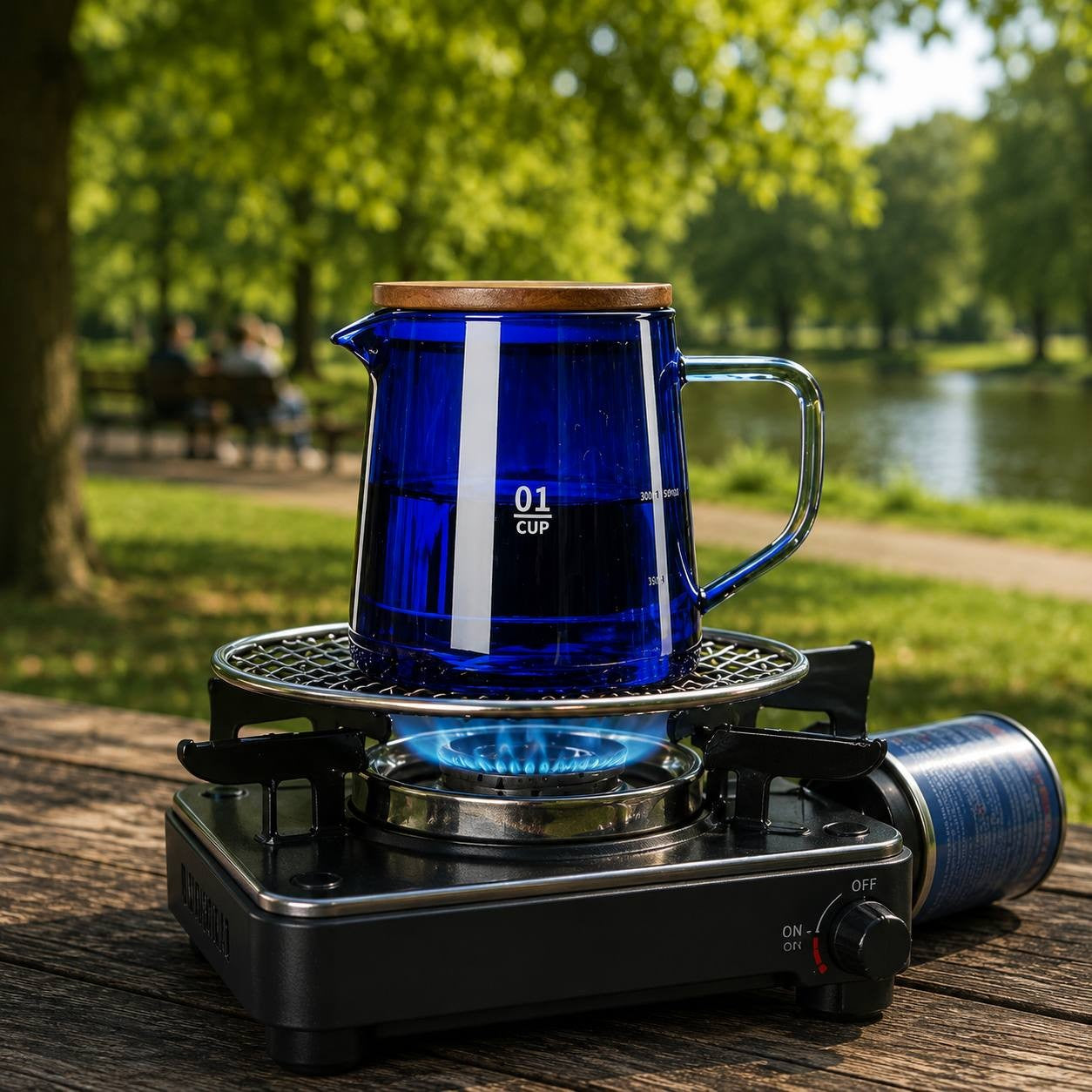Cobalt blue glass pour-over coffee kettle with a wooden lid heating on a portable gas stove outdoors on a wooden park table.