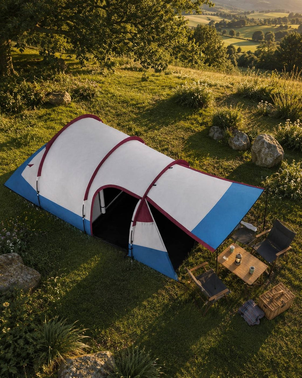 Aerial view of a blue, white, and burgundy camping tent pitched on a grassy hillside in a sunny rural valley.