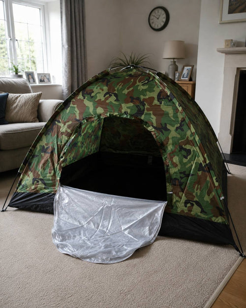 A dome-shaped camouflage camping tent set up on a carpet inside a bright living room next to a sofa and window.