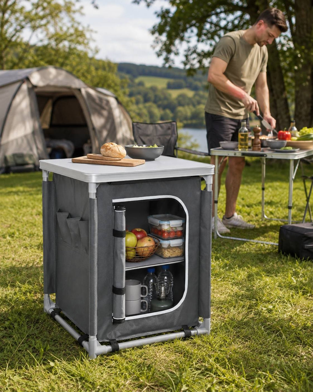 Grey camping kitchen cupboard set up on grass near a tent and a person cooking at a table in the background.