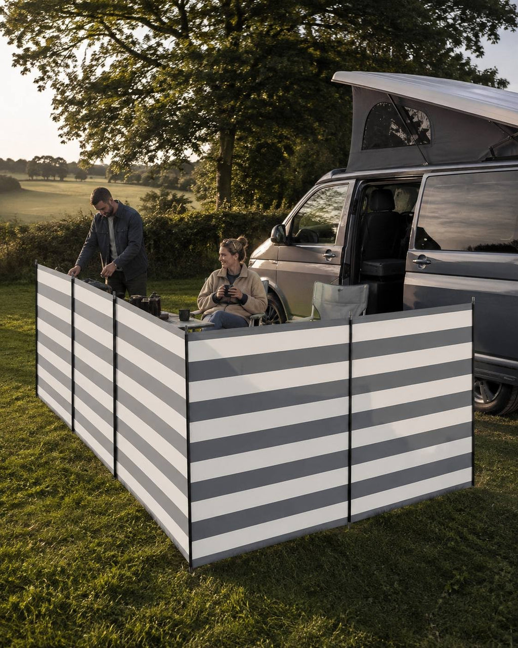 Grey-and-white striped camping windbreak enclosing a camping setup next to a campervan on a grassy field at sunset, with a man standing near a camp kitchen and a woman sitting holding a mug.