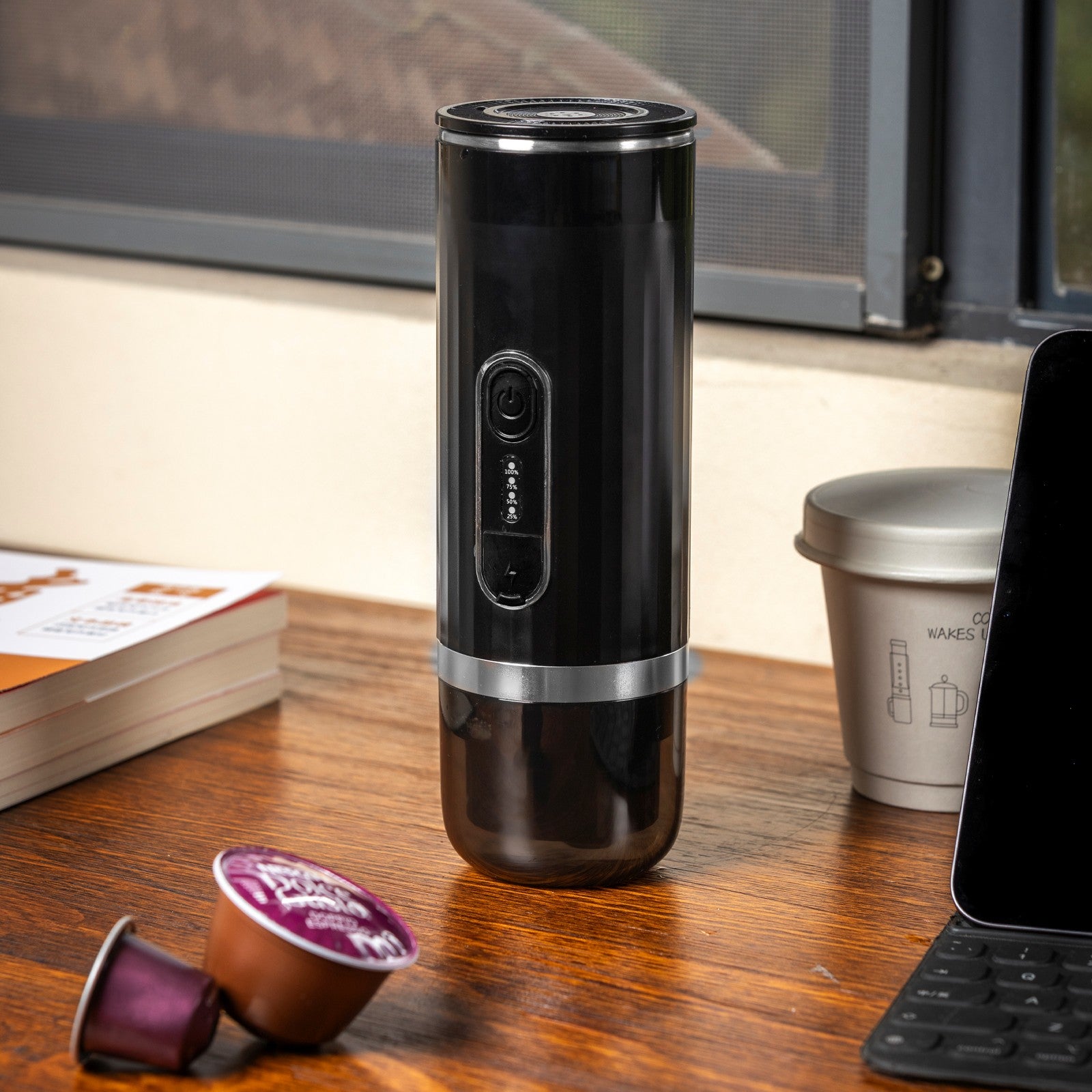 Black capsule portable espresso machine standing on a wooden desk next to coffee pods and a tablet