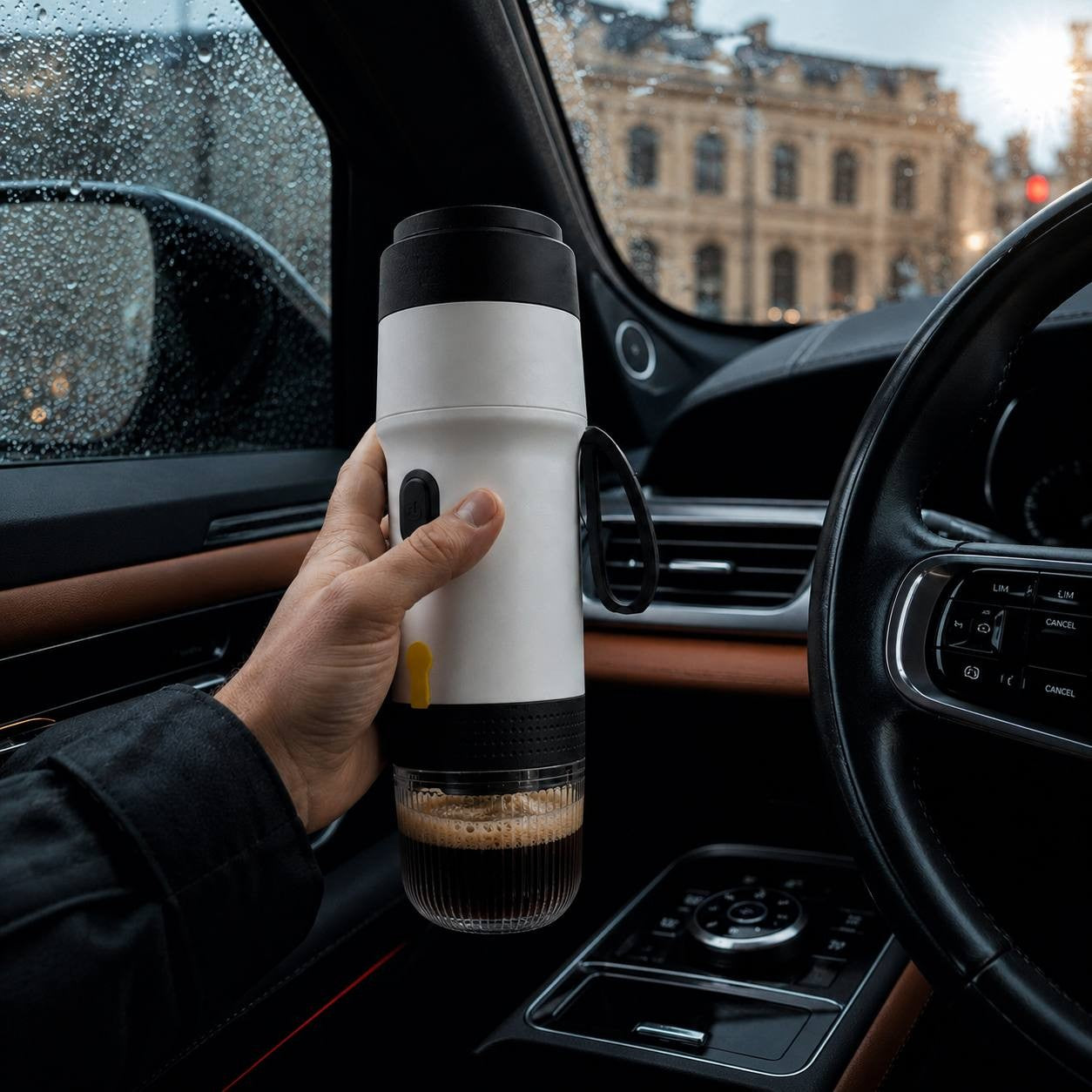 A driver's hand holding a white portable electric espresso machine inside a luxury car with raindrops on the window and a city building background.