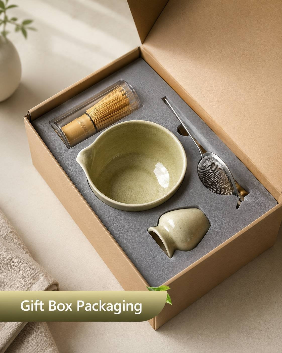 Matcha tea set in gift box packaging, including ceramic bowl, bamboo whisk in a clear case, strainer, and scoop on gray foam insert.