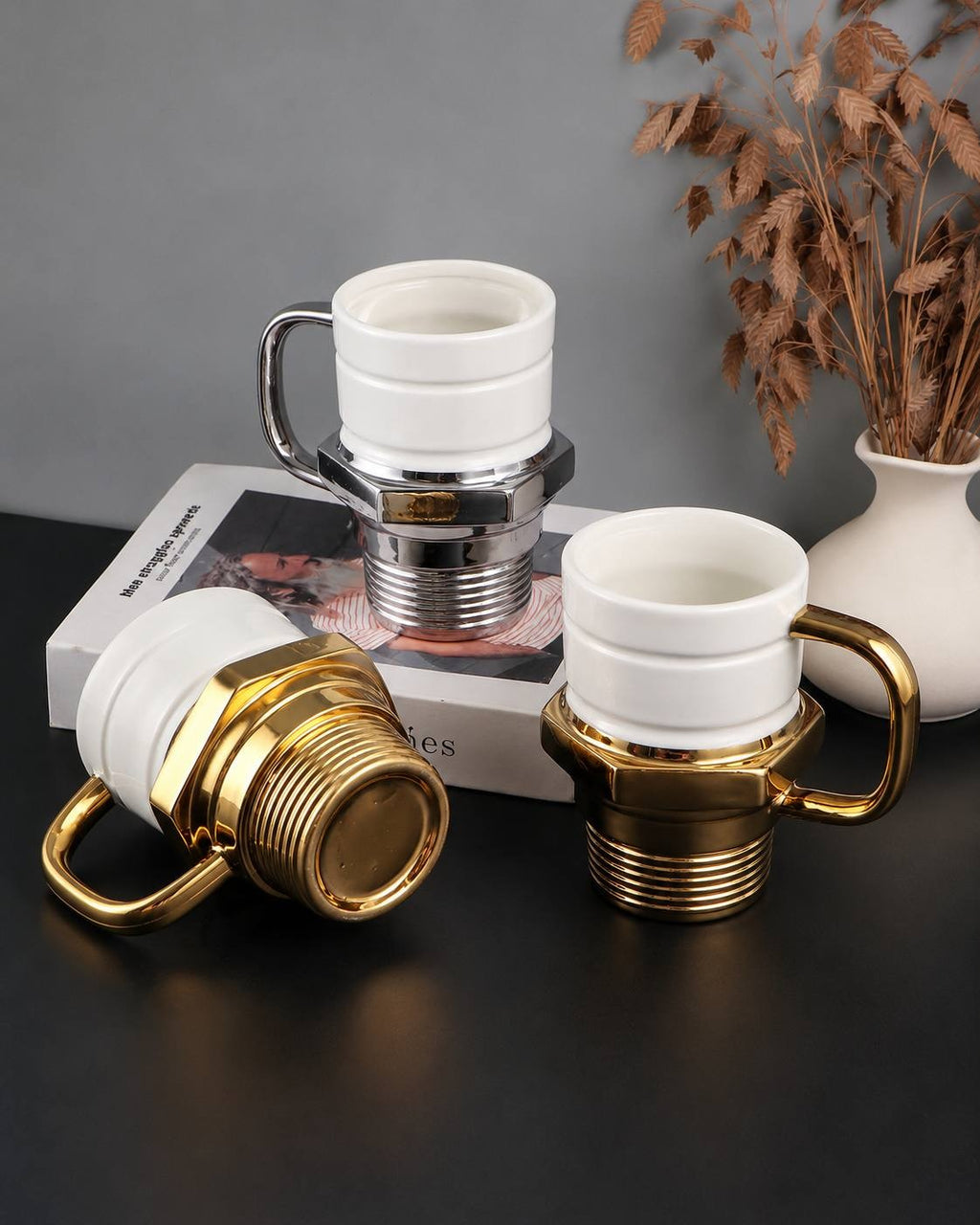 A stylish product shot featuring a set of two spark plug-shaped ceramic mugs, one in gold and one in silver, arranged on a dark table with a book and a vase of dried flowers, with one mug lying on its side to show the threaded bottom.