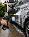 A close-up action shot of a person using a 24V cordless pressure washer equipped with a soap foam bottle to spray thick cleaning foam onto the side of a grey