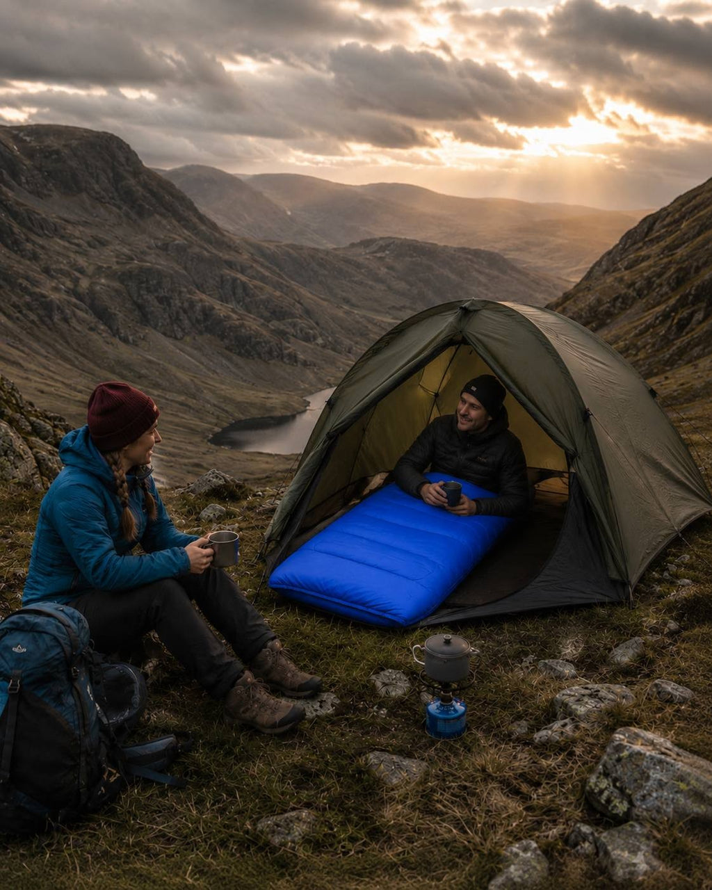 Woman sitting outside a tent on a mountain slope holding a mug while a man sits inside the tent resting on a blue sleeping bag during sunset.