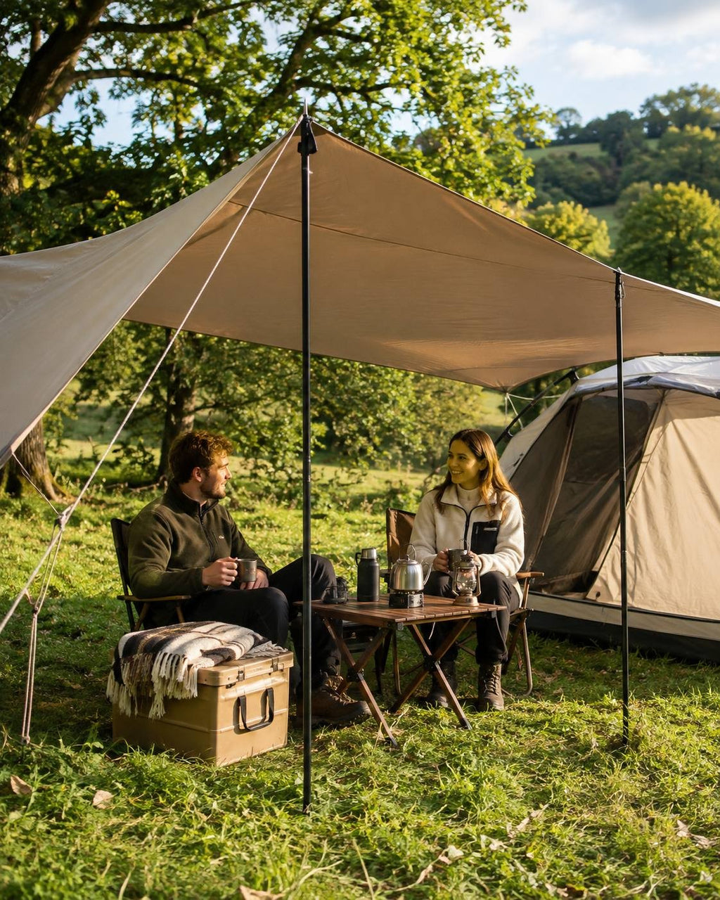 A couple camping in a lush green field, sitting under a beige tarp shelter next to a tent, enjoying coffee from mugs.