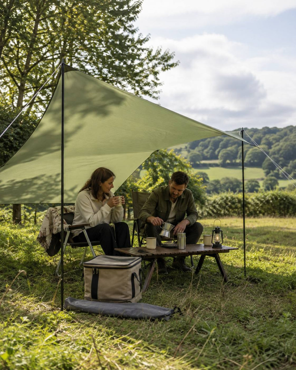A couple sitting under an olive green camping tarp shelter in a field, with a man pouring coffee from a moka pot.