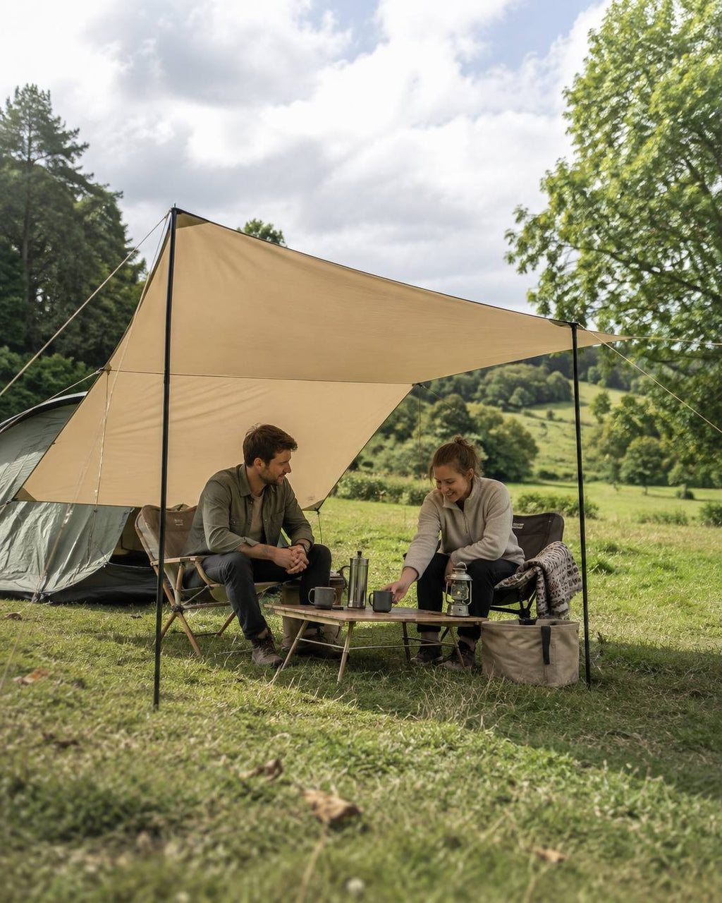 A man and woman relaxing together under a beige camping tarp shelter set up in a grassy field with trees in the background.