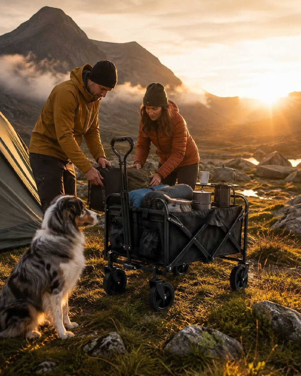 Couple unpacking camping gear and sleeping bags from a black folding utility wagon next to a tent in the mountains at sunset with a dog.