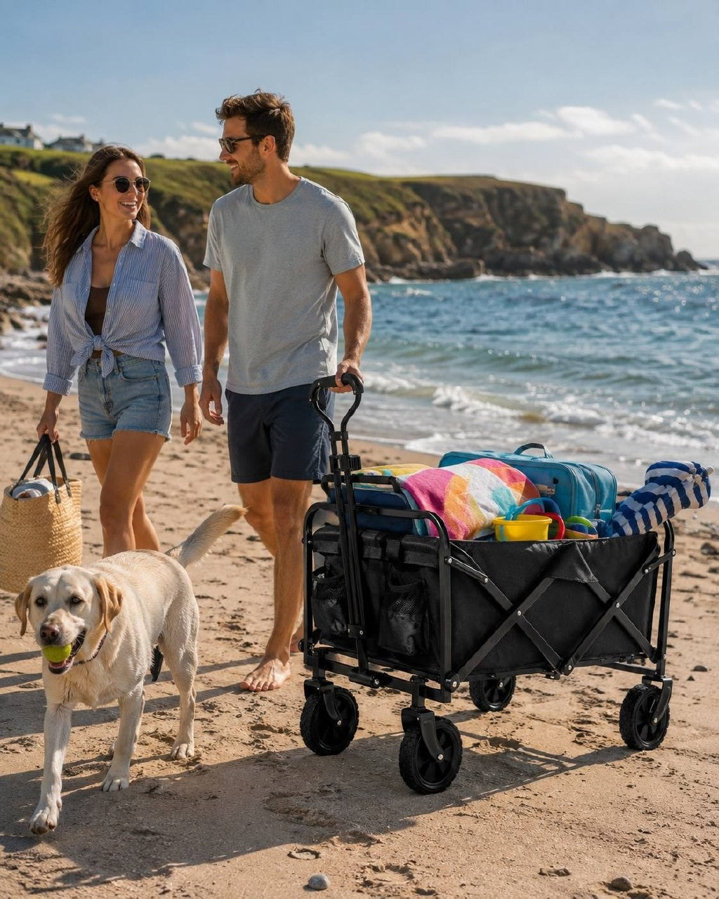 Couple walking along a sandy beach with a dog holding a ball and a black utility wagon filled with towels and bags near the sea.