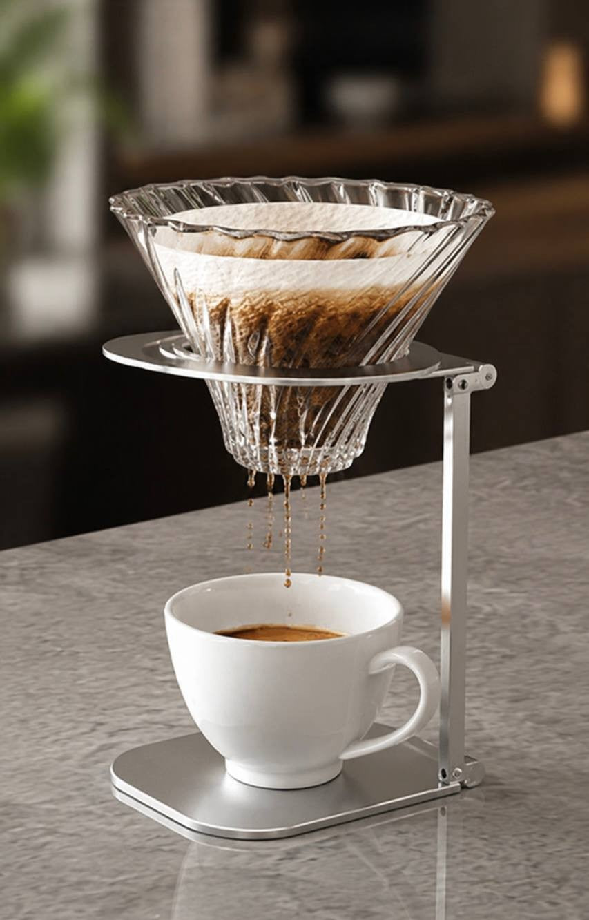 Clear glass V60 coffee dripper mounted on a sleek silver metal stand brewing pour-over coffee into a white ceramic cup