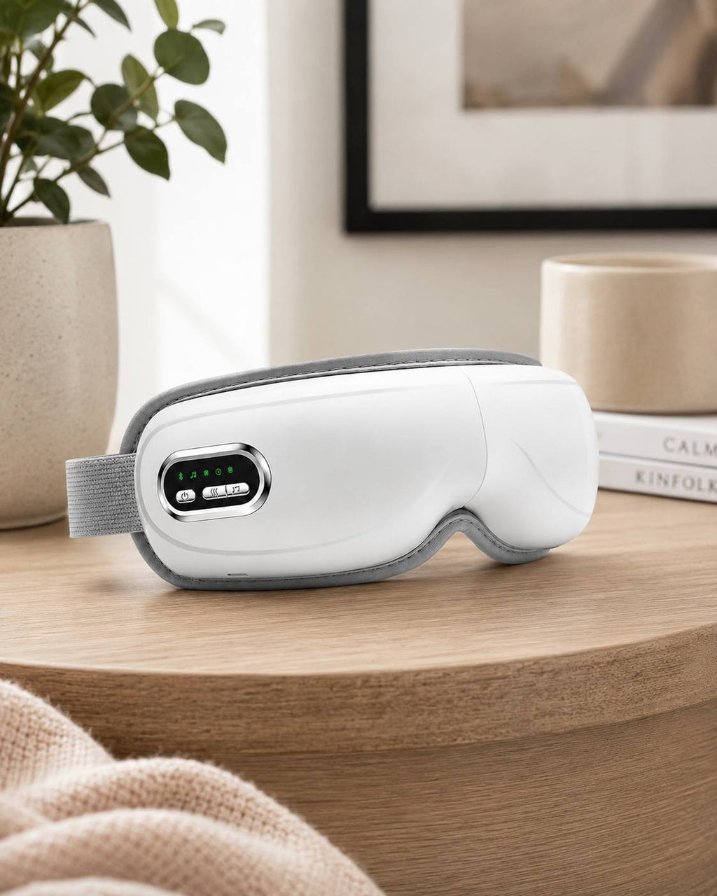 A white electric airbag eye massager placed on a wooden tabletop in a cozy home setting with a plant and decor in the background.
