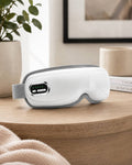 A white electric airbag eye massager placed on a wooden tabletop in a cozy home setting with a plant and decor in the background.
