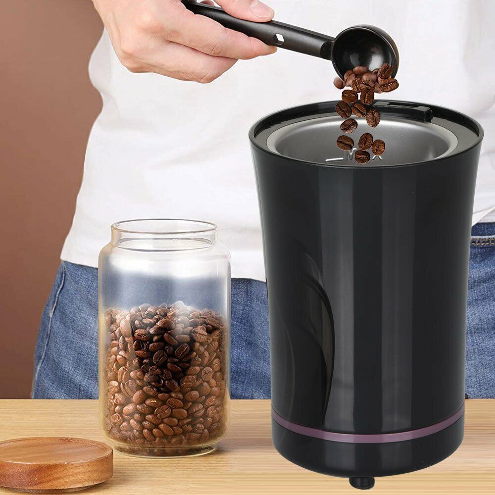 Person pouring coffee beans from a scoop into a black electric grinder next to a glass jar of beans.