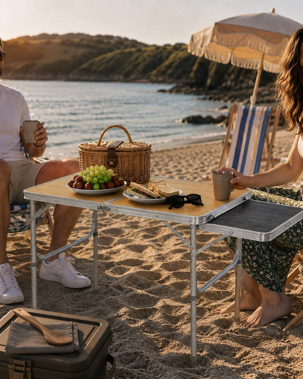 Couple sitting in beach chairs at a folding camping table with mugs and sandwiches, with the ocean in the background.