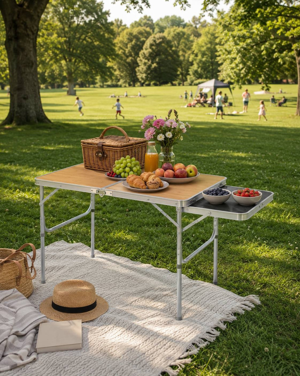 Wooden folding camping table set up on a picnic blanket in a green park with a wicker basket, fruit, and people relaxing in the background.