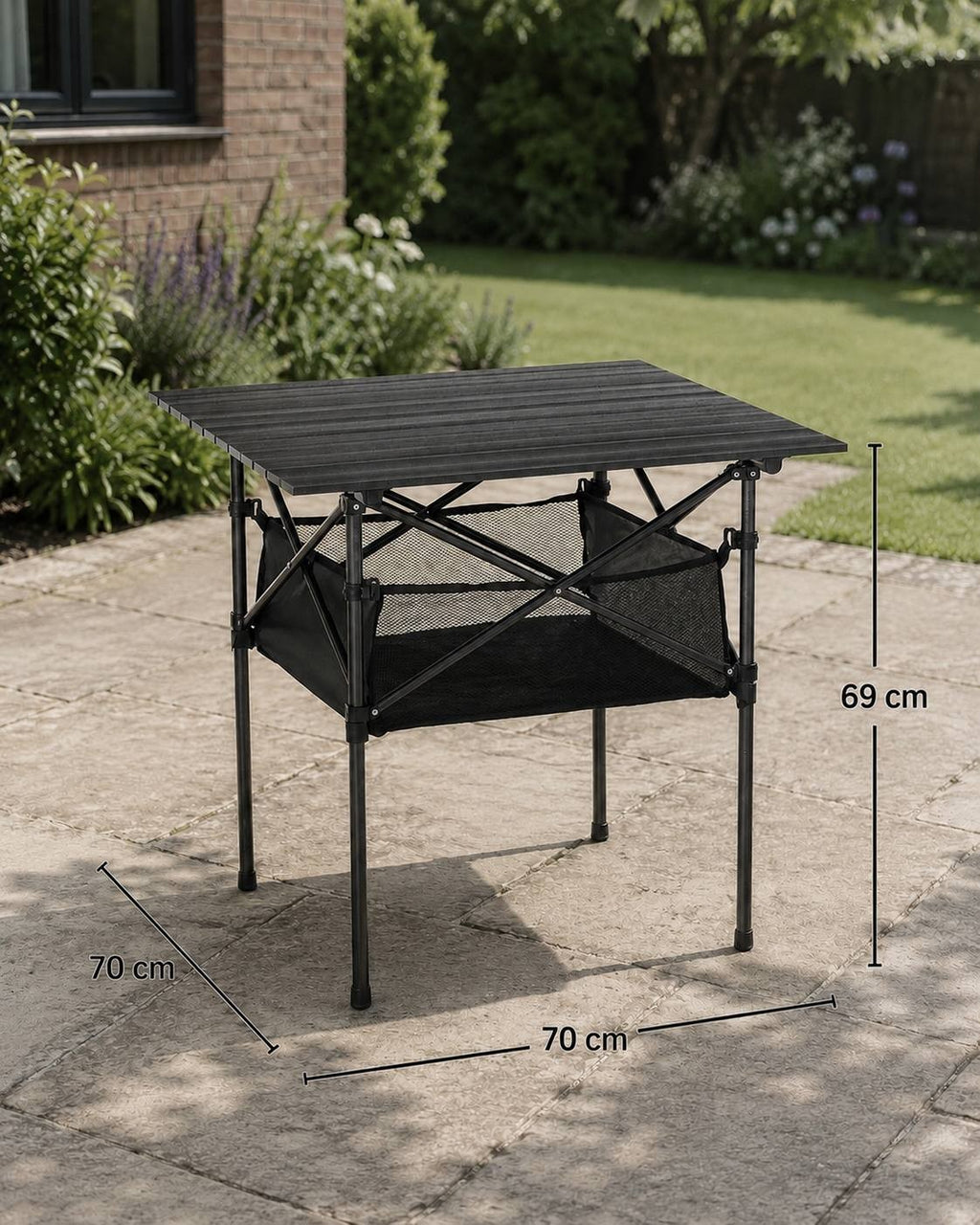A black folding aluminum camping table with a slatted top and mesh storage net set up on a stone patio in a backyard, featuring dimension overlays of 70 cm width, 70 cm length, and 69 cm height.