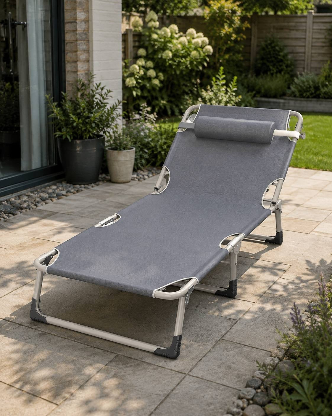 A grey folding lounge bed with a headrest pillow set up on a stone patio tile floor outside a house with green plants in the background.