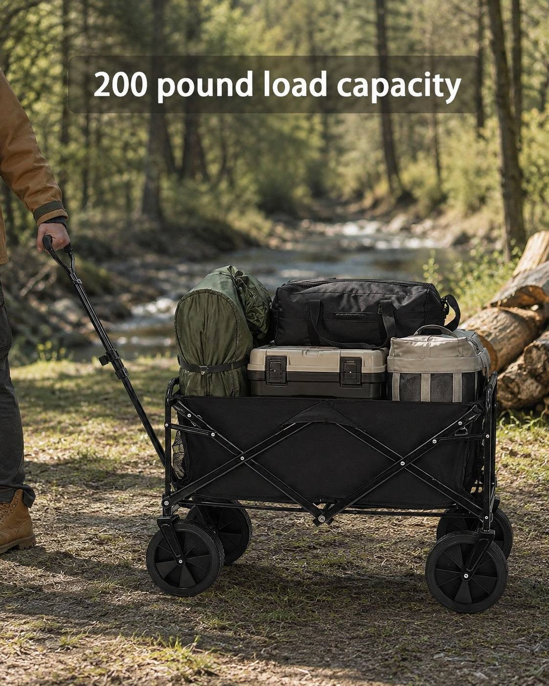 Black folding utility wagon loaded with camping bags, a tent, and gear on a trail near a forest stream with a person holding the handle.
