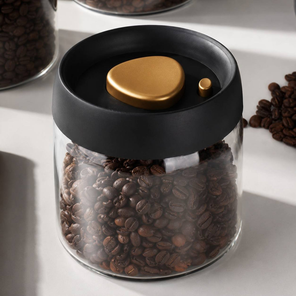 Glass coffee bean storage canister with a black and gold vacuum lid sitting on a white table.
