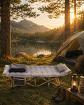 Grey folding camping cot with a quilted mattress, blanket, and pillow set up on grass next to a tent by a lake at sunset.
