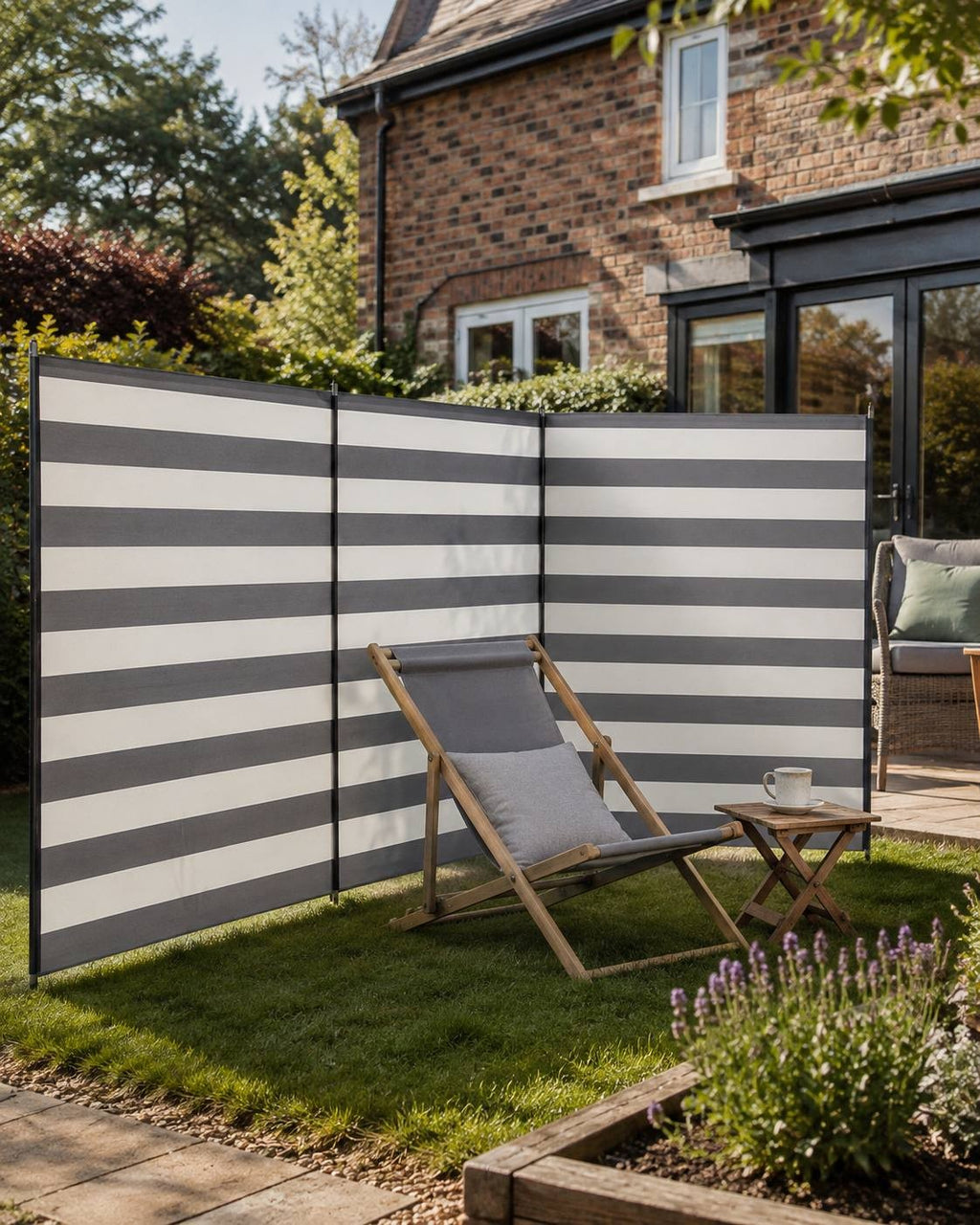 Striped grey and white multi-panel camping windbreak set up on a green lawn in a backyard next to a brick house, featuring a wooden deckchair and a small table with a mug.