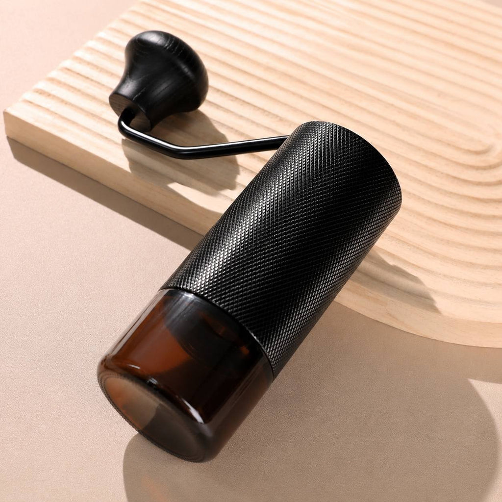 Close-up of a handheld manual coffee mill lying on a wavy wooden tray showcasing the amber glass grounds catcher.