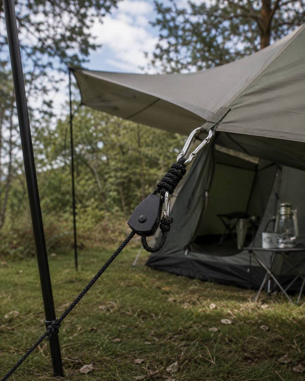 A detailed view of a black rope ratchet hanger hooked to a tarp corner loop and a support pole in a wooded campsite.