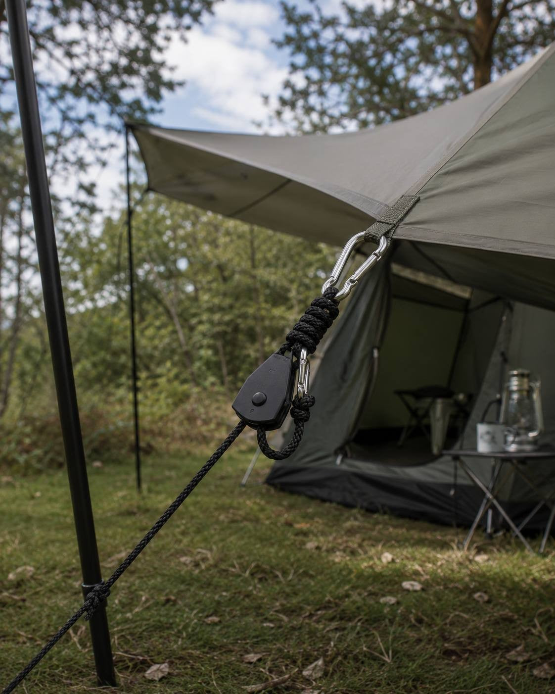 A detailed view of a black rope ratchet hanger hooked to a tarp corner loop and a support pole in a wooded campsite.