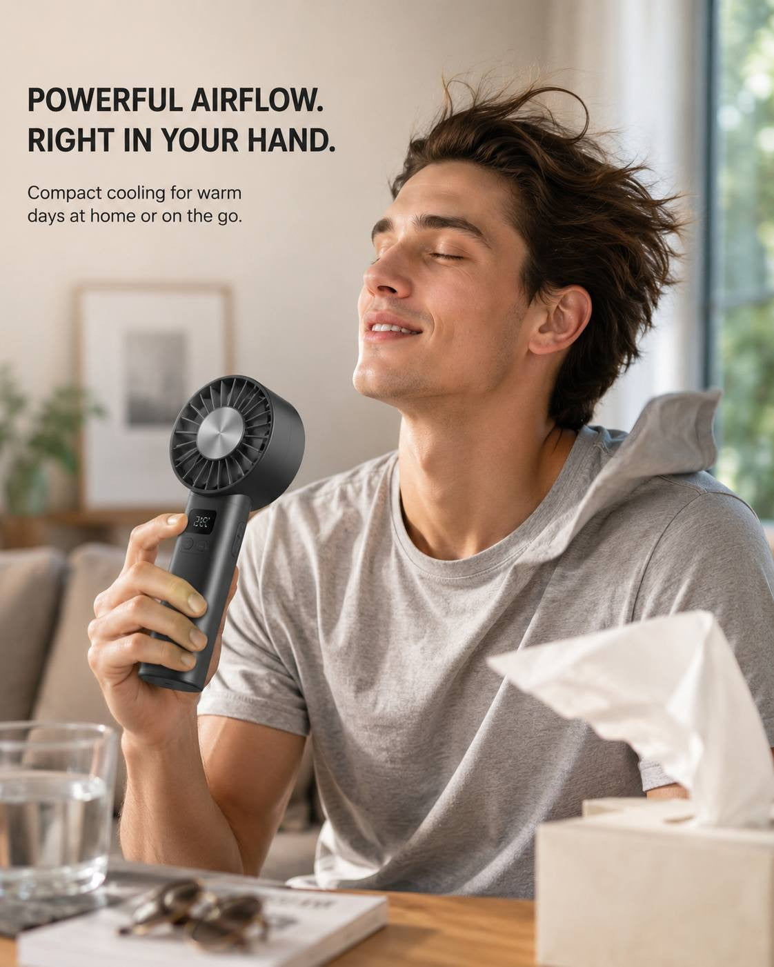 A man relaxing indoors in a grey t-shirt, holding a black portable handheld cooling fan and enjoying the airflow on a warm day.