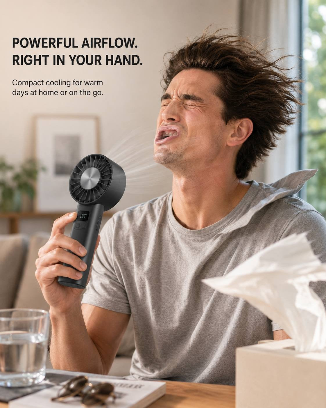 A man sitting indoors in a grey t-shirt, making a funny expression as he tests the powerful airflow of a black portable handheld cooling fan.
