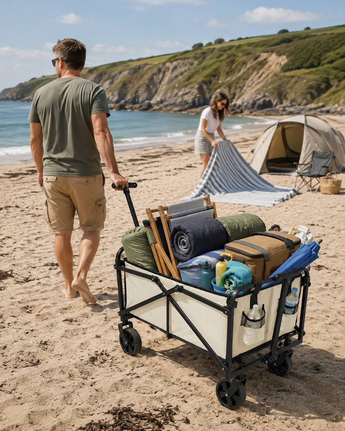 Man pulling a beige and black folding utility wagon filled with camping gear, sleeping bags, and a cooler across a sandy beach, with a woman unpacking a blanket near a tent in the background.