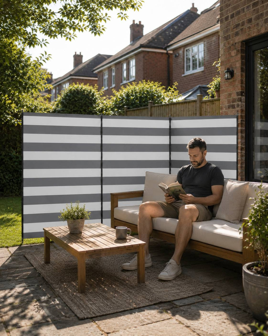 Man sitting on an outdoor sofa reading a book next to a grey-and-white striped windbreak on a patio with a brick house in the background.