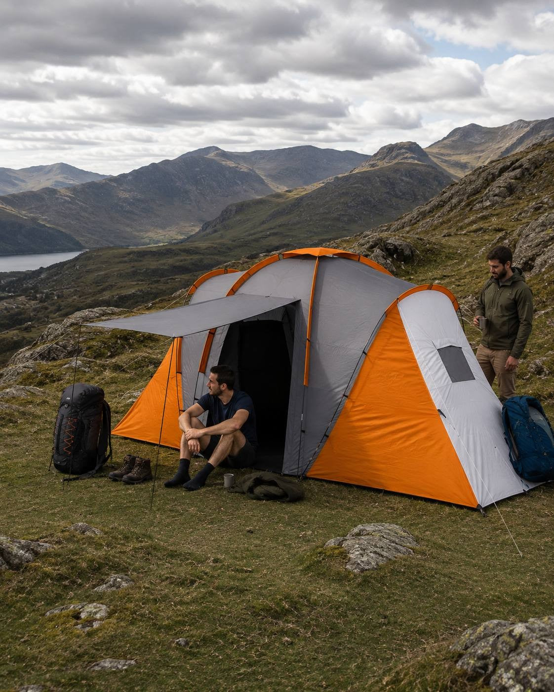 Man sitting by the entrance of a large orange and grey camping tent set up on a grassy mountain slope overlooking a lake and distant mountains.