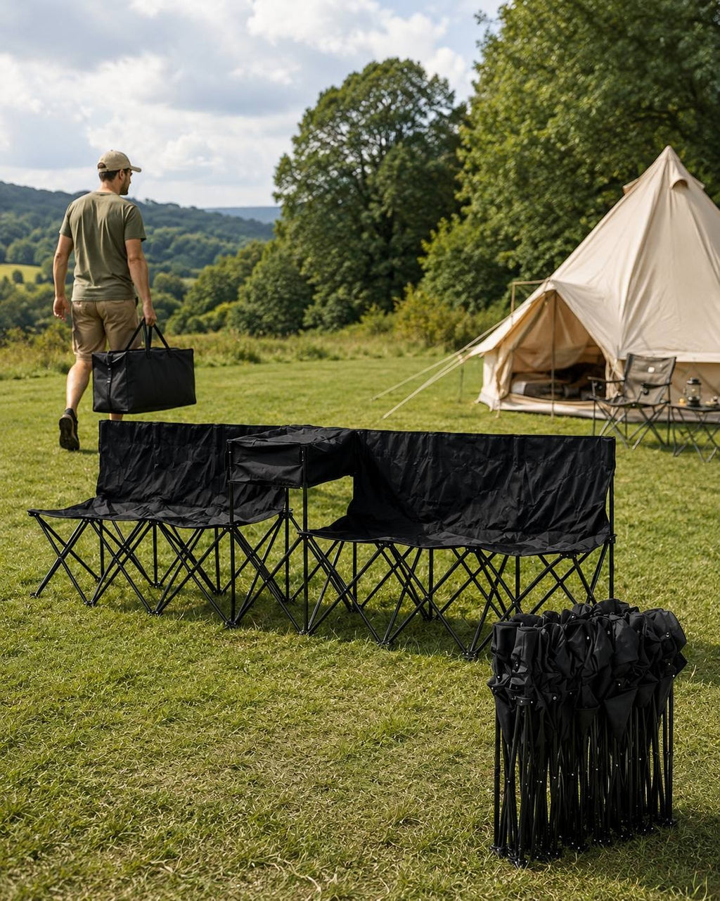 Man in a cap and green t-shirt carrying a black duffel bag across a grassy field past a set of black folding outdoor benches and a bell tent.