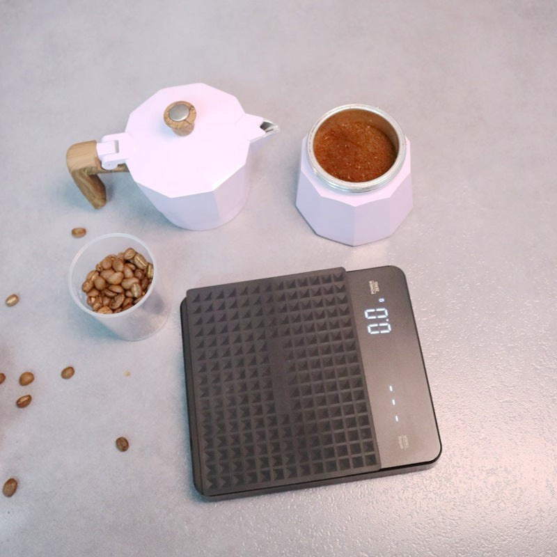 Digital kitchen scale with coffee beans, a white teapot, and a container of ground coffee on a light gray surface.